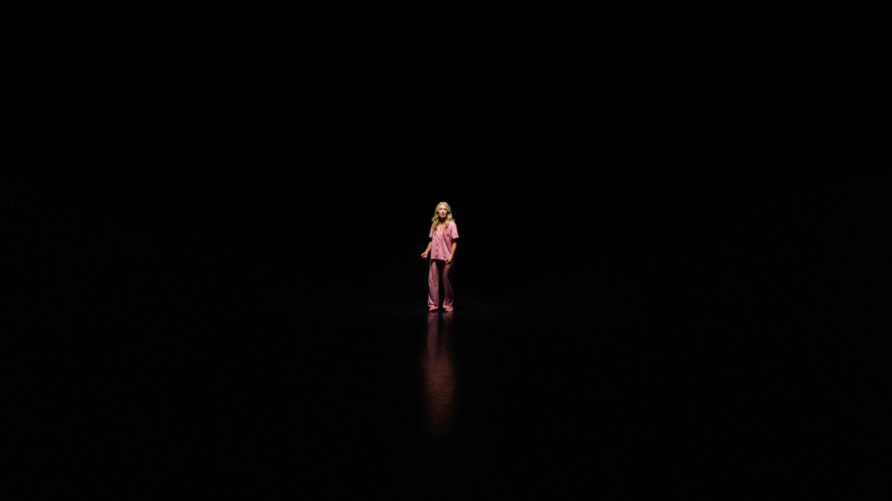 A woman in pink pajamas stands alone in a dark space under a narrow spotlight.